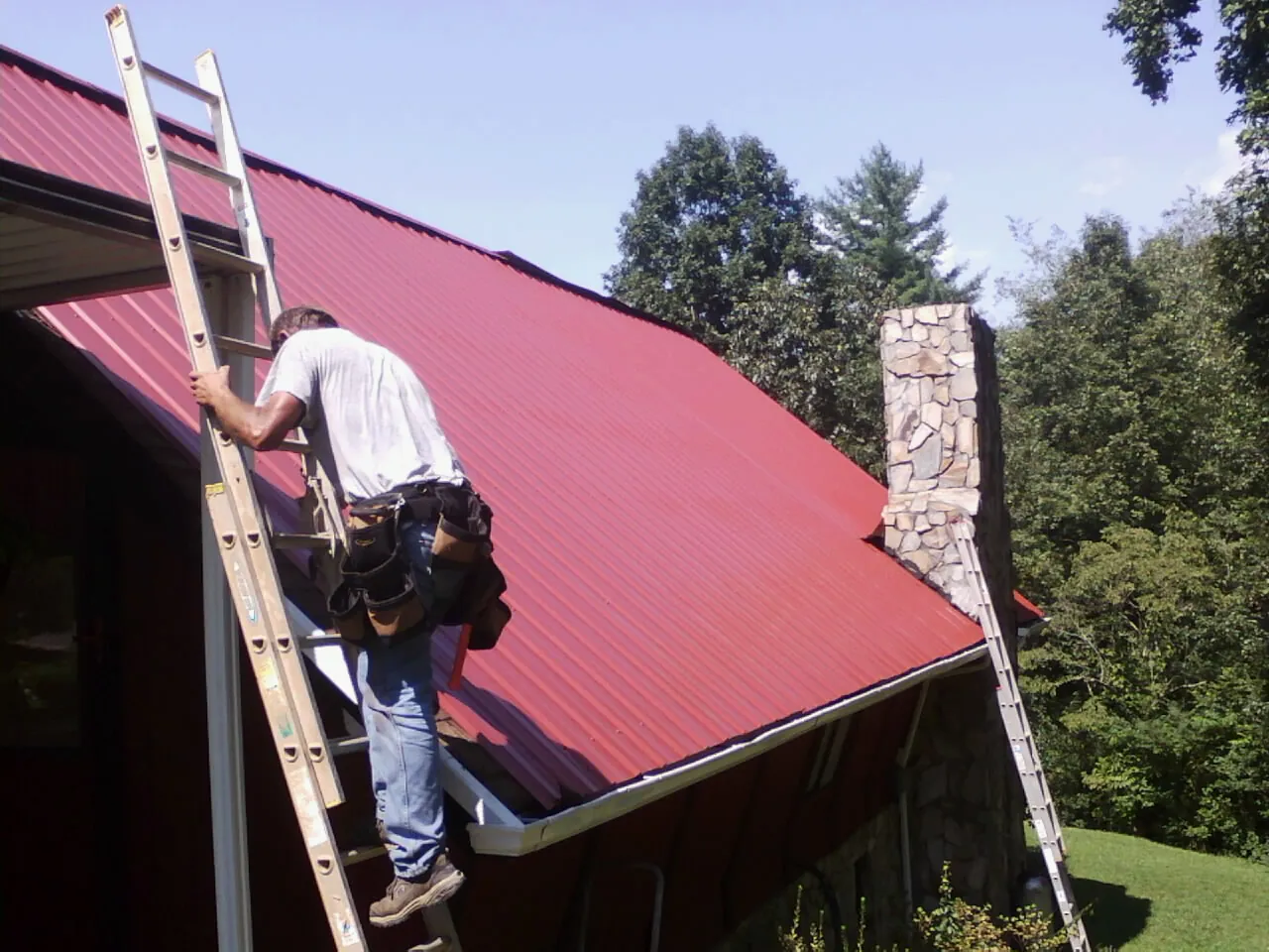 Roofer on a ladder beside a red metal roof and stone chimney — Soffit & Fascia Repair by Roofing Brothers in Sandersville